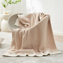Beige knitted throw blanket draped over couch arm, with cream border and soft fringed edge.