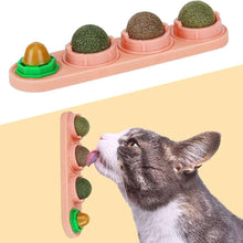 Pink cat wall treat toy with green-brown catnip balls; gray cat reaching to lick.