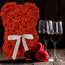 Red artificial rose bear with white ribbon bow beside red roses and wine glasses